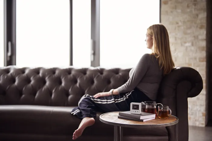 The future of smart heating - A woman sits on a sofa looking into the distance, next to her is a table with a smart thermostat, a book, and a hot drink