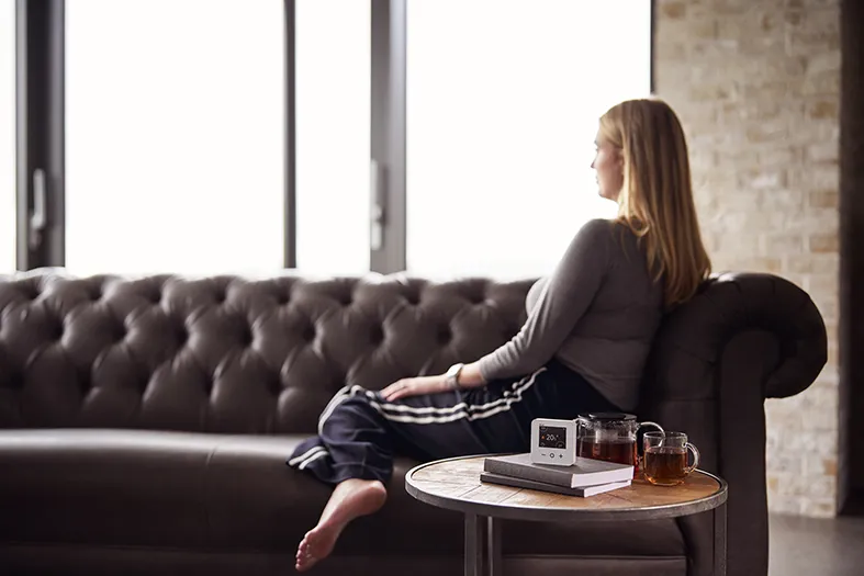 The future of smart heating - A woman sits on a sofa looking into the distance, next to her is a table with a smart thermostat, a book, and a hot drink